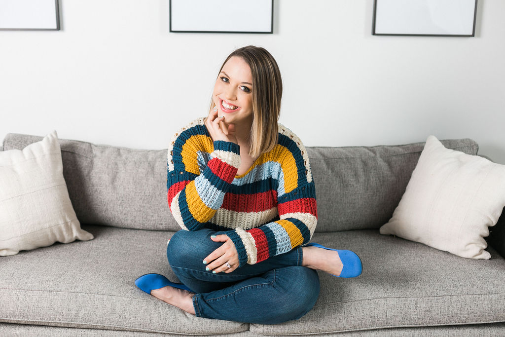 Emilie Steinmann is wearing a bold colored sweater and jeans and is sitting cross legged on a gray sofa.